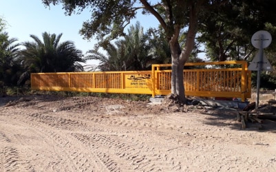 שער קונזולי גדול מברזל בצבע צהוב מרחף מעל שטח חולי ולוח במרכזו עם חריטת לייזר, ניתן לראות עצים ברקע