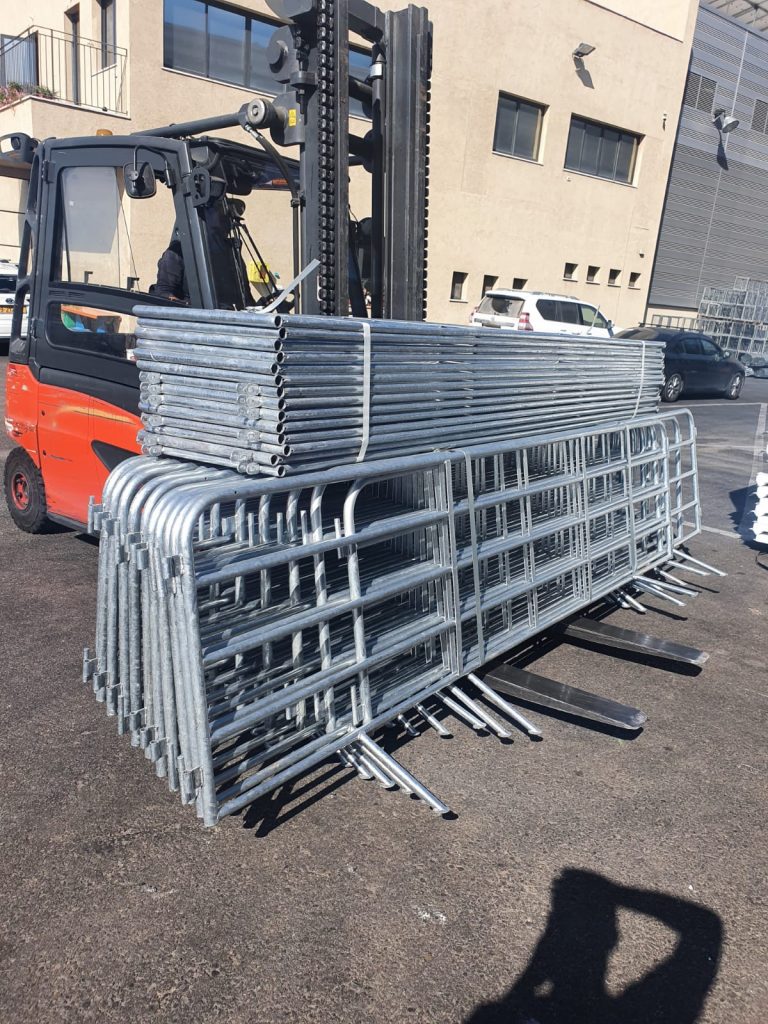 מלגזה כתומה נושאת ערימה גדולה של מסגרות מתכת מגולוונות. המסגרות כוללות צינורות עגולים המחוברים בצורת גדרות.