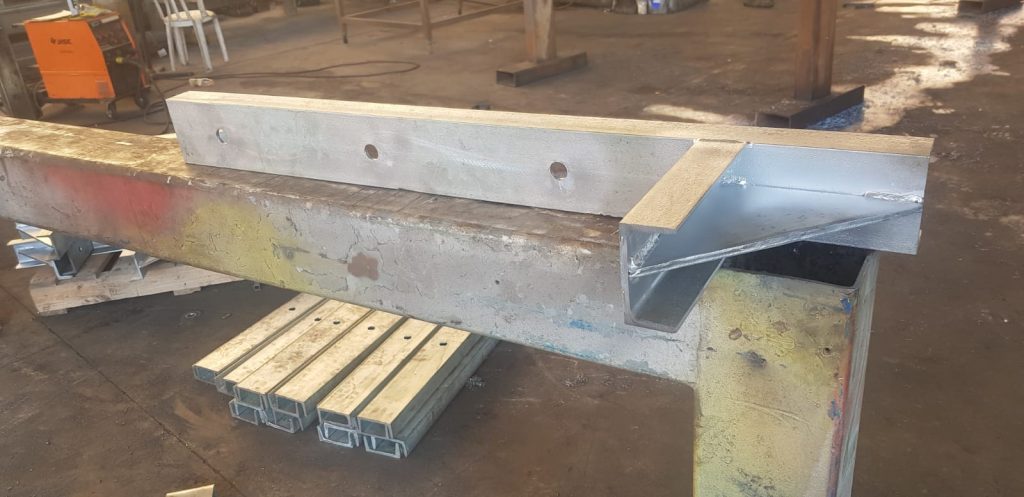 חלק מתכת מגולוון עם זווית מרותכת, מונח על קורה מתכתית. בחלקו של החלק נראים שני חורים גדולים. ברקע ניתן לראות אזור עבודה תעשייתי עם מכונות וכלים