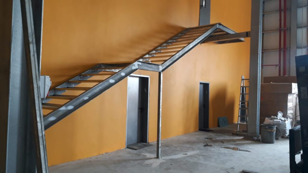 גרם מדרגות מתכת המחובר לקיר בצבע כתום במבנה תעשייתי. המדרגות מובילות לקומה עליונה ויש להן מבנה פתוח ללא מעקה. מתחת למדרגות ישנן שתי דלתות מתכתיות,