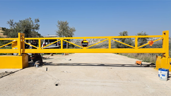 שער קונזולי ממתכת צהוב עם דפוס צולב מותקן על דרך. על השער מופיע לוגו "פנחס היישריק בע"מ". אדם עובד ליד בסיס השער, וברקע נראים עצים ונוף הררי.