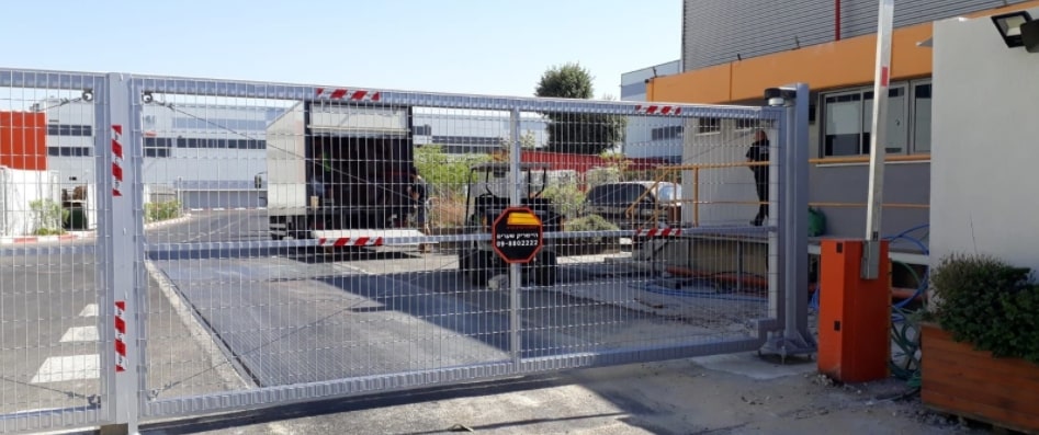 שער רשת מתכתית בגוון אפור בהיר בכניסה למתחם, על רקע מבנה תעשייתי צבוע בצבעים שונים