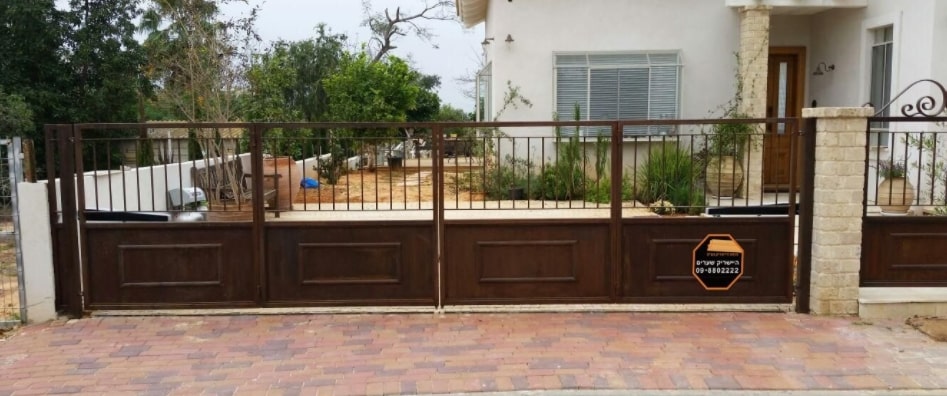 שער מתכת בגוון חום עם פאנלים דמויי עץ ודפנות עם סורגים, המוביל לחצר בית פרטי.