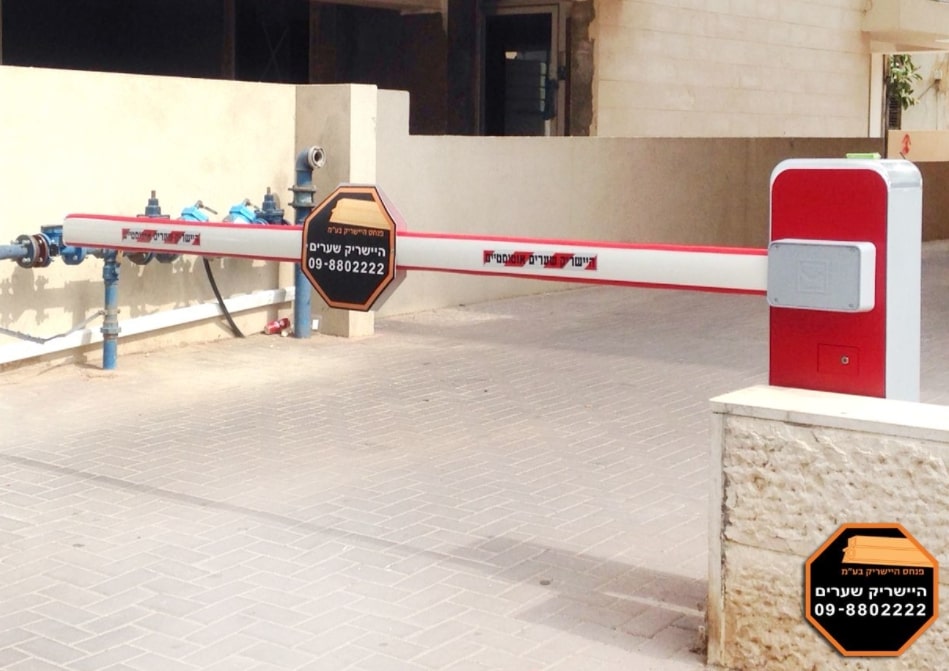 מחסום חשמלי בצבע לבן ואדום בכניסה לחניון מגורים ניתן לראות על המוט עצמו תמרורי אזהרה ושלט מתומן של לוגו היישריק שערים -09-8802222